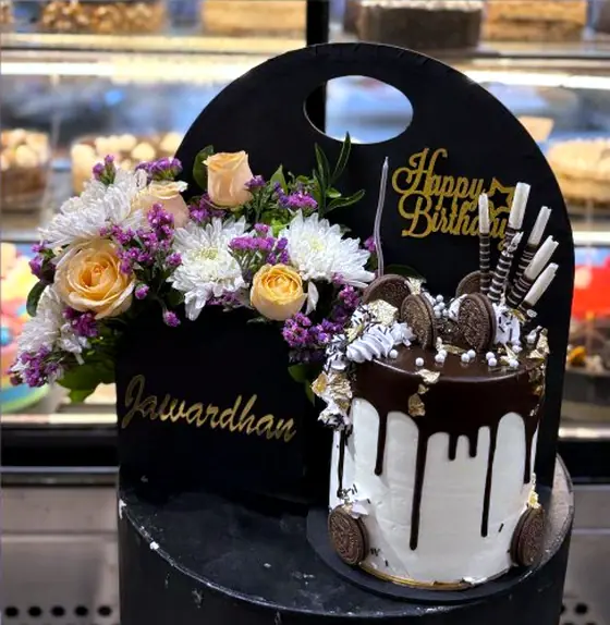 A drip cake with Oreo biscuits beside a small carry-box of fresh peach and white flowers, both branded Brother's Bakers